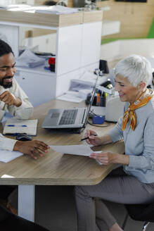 Glückliche ältere Frau, die mit einem Verkäufer über ein Dokument am Schreibtisch diskutiert - IKF01633