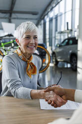 Happy customer shaking hands with salesperson at desk in car showroom - IKF01642