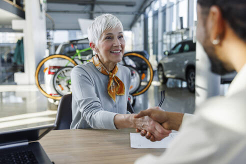 Happy senior woman shaking hands with salesperson at desk in car showroom - IKF01643