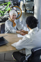 Happy woman shaking hands with salesperson at desk in car showroom - IKF01644