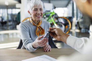 Salesperson giving car keys to smiling woman sitting at desk - IKF01652