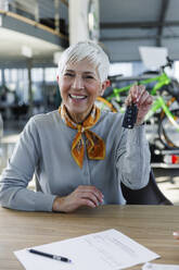 Happy senior woman holding car keys at desk - IKF01655