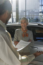 Glückliche ältere Frau mit Verkäuferin, die ein Dokument hält und ein Auto im Ausstellungsraum zeigt - IKF01664