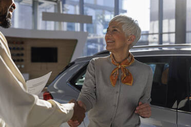 Happy senior woman shaking hands with salesperson near car at showroom - IKF01665
