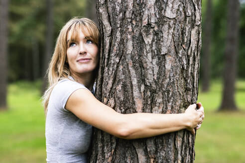 Lächelnde blonde Frau, die einen Baum im Wald umarmt - WPEF08284