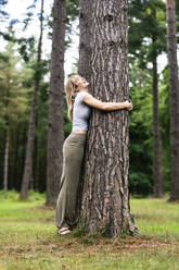 Cheerful young beautiful woman hugging tree in forest - WPEF08286