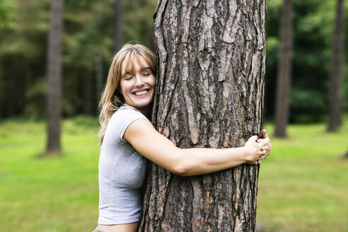 Lächelnde junge schöne Frau, die einen Baum im Wald umarmt - WPEF08288