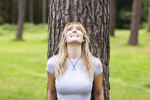 Happy beautiful blond woman leaning on tree - WPEF08294