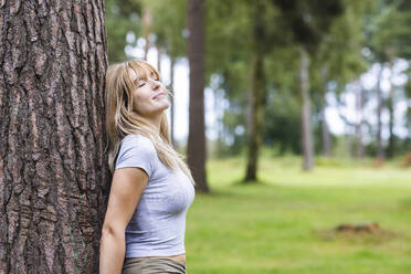 Smiling beautiful woman leaning on tree in forest - WPEF08295
