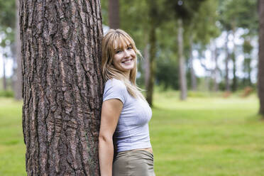 Happy young beautiful woman leaning on tree in forest - WPEF08298