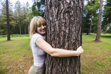 Smiling young woman hugging tree trunk in forest - WPEF08299