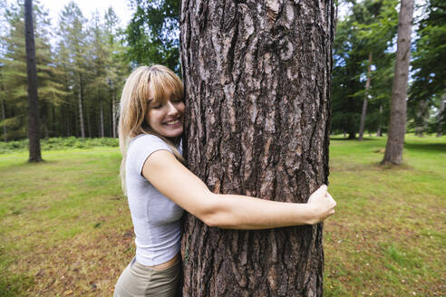Lächelnde junge Frau, die einen Baumstamm im Wald umarmt - WPEF08299