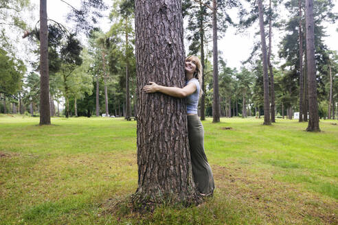 Lächelnde junge Frau, die einen Baum im Wald umarmt - WPEF08300