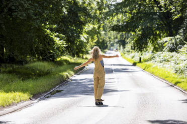Young woman with arms outstretched skateboarding in forest on sunny day - WPEF08323