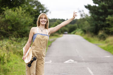 Happy beautiful woman holding skateboard and hailing ride on road - WPEF08329