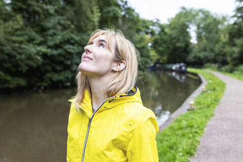 Smiling woman wearing yellow raincoat standing near trees - WPEF08380