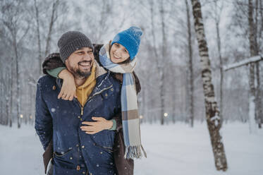 Glücklicher Mann, der eine Frau im Winter im Park huckepack nimmt - ANAF02704