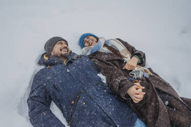 Verspieltes reifes Paar hält sich an den Händen und liegt im Schnee im Park - ANAF02705