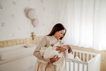 Happy mother holding baby girl in arms near crib at home - EBBF08376