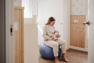 Mother sitting on fitness ball and holding daughter at home - EBBF08377