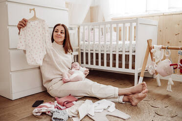 Woman holding daughter and choosing baby clothes near crib - EBBF08402