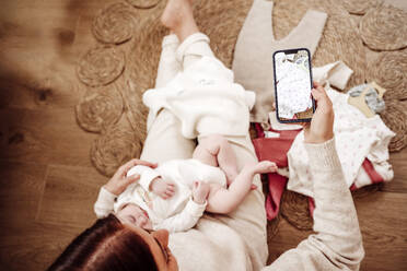 Frau hält Tochter und fotografiert Babykleidung mit dem Smartphone - EBBF08403