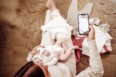 Woman holding daughter and photographing baby clothes through smart phone - EBBF08403