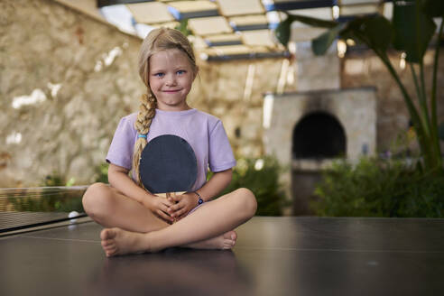 Lächelndes Mädchen mit Tischtennisschläger im Schneidersitz in einer Villa - ANNF00823