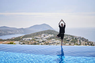 Man practicing tree pose at the edge of swimming pool at villa - ANNF00827