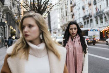 Serious woman wearing scarf and standing with friend on sidewalk in city - PBTF00431