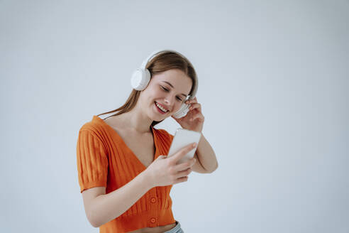 Lächelnde Frau, die drahtlose Kopfhörer trägt und ein Smartphone vor einem weißen Hintergrund benutzt - NLAF00237