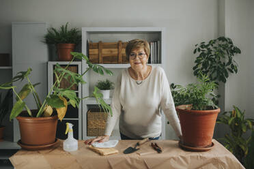 Smiling elderly woman leaning on table with plants at home - DMGF01174