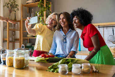 Glückliche multirassische Frauen, die ein Selfie mit ihrem Handy in der Küche zu Hause machen - OIPF03899