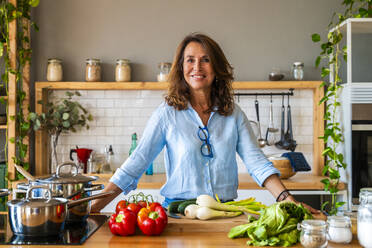 Smiling senior woman standing by vegetable on kitchen counter at home - OIPF03912