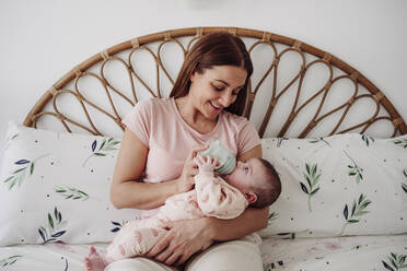 Smiling mother feeding milk to daughter on bed at home - EBBF08416