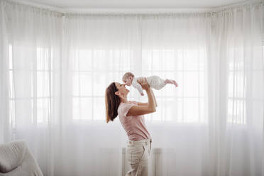 Glückliche Frau spielt mit ihrer Tochter vor einem Fenster zu Hause - EBBF08424