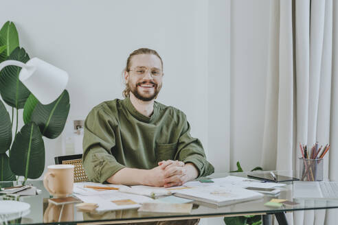 Smiling furniture designer sitting with hands clasped at desk in office - YTF01716