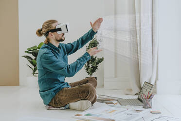 Interior designer wearing virtual reality simulator and sitting on floor with laptop at office - YTF01732