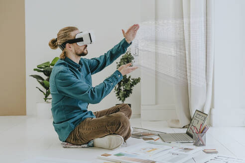 Interior designer wearing virtual reality simulator and sitting on floor with laptop at office - YTF01732