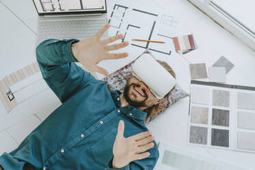 Smiling interior designer wearing VR glasses and lying on floor at office - YTF01737