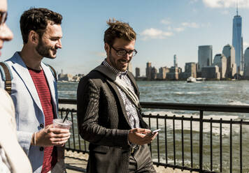 Happy businessmen with smart phone walking on promenade in city - UUF31153