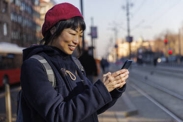 Lächelnde reife Frau, die ein Smartphone an einer Straßenbahnhaltestelle benutzt - IKF01710