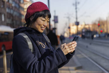Smiling mature woman using smart phone at tram station - IKF01710