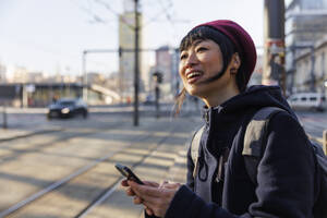 Mature woman standing with smart phone at tram station - IKF01712