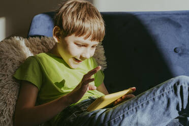 Excited boy using smart phone sitting on sofa at home - OSF02381