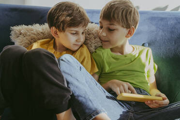 Siblings sharing smart phone sitting on sofa at home - OSF02382