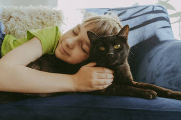 Smiling boy with eyes closed lying down with cat on sofa at home - OSF02384