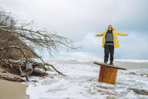 Unbekümmerte Frau steht mit ausgestreckten Armen auf einer Holzspule am Strand - OLRF00138