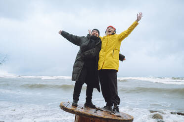 Fröhliche Freunde stehen mit ausgestreckten Armen auf einer Holzspule am Strand - OLRF00139