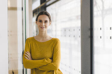 Smiling young businesswoman standing with arms crossed in corridor - JOSEF23363